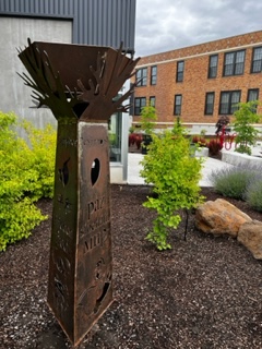 A tall metal sculpture placed among the Ginkgo trees in a planting bed with the school buildings in the background
